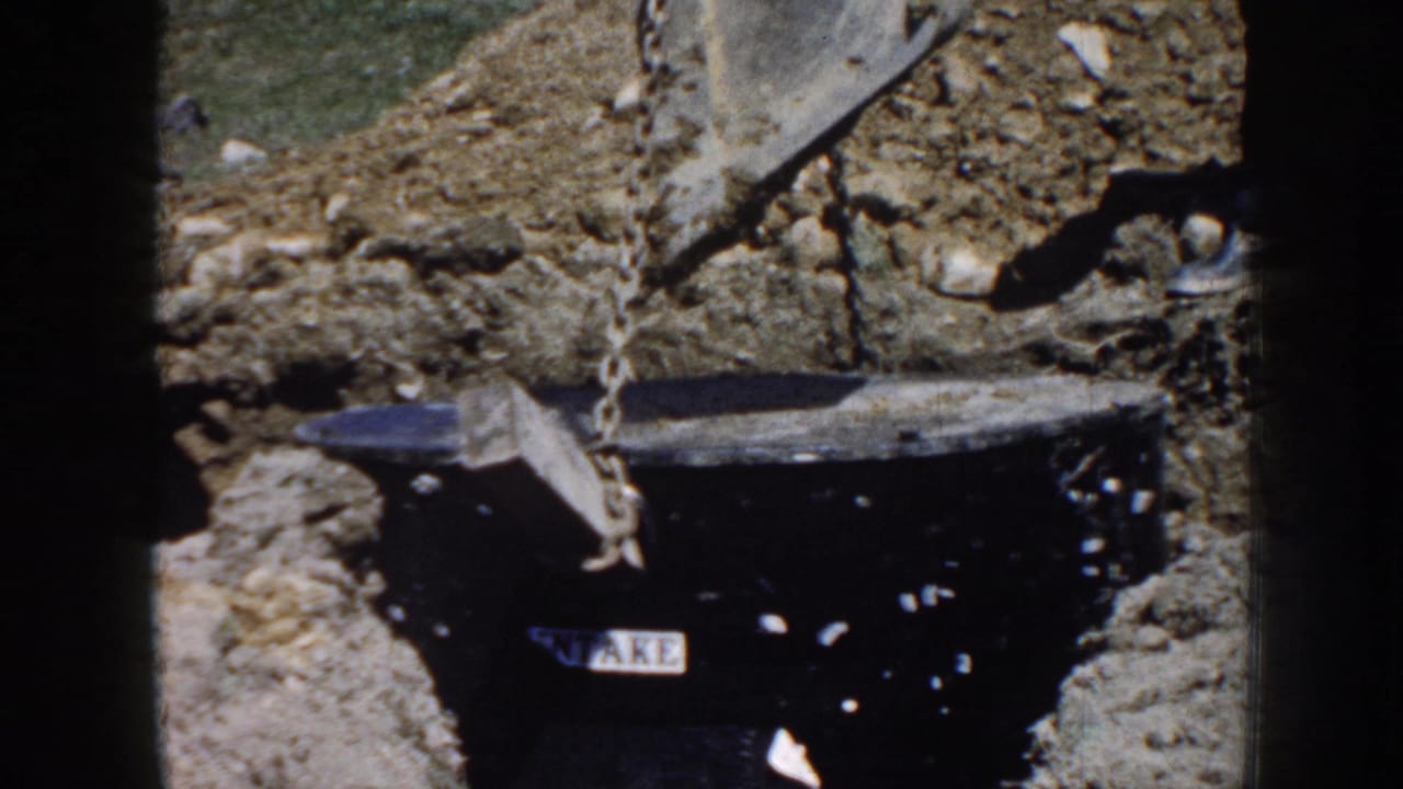 1958 Nebraska 8mm frame showing a black utility-like cylinder in a dug trench, suspended by a chain from an excavator bucket.