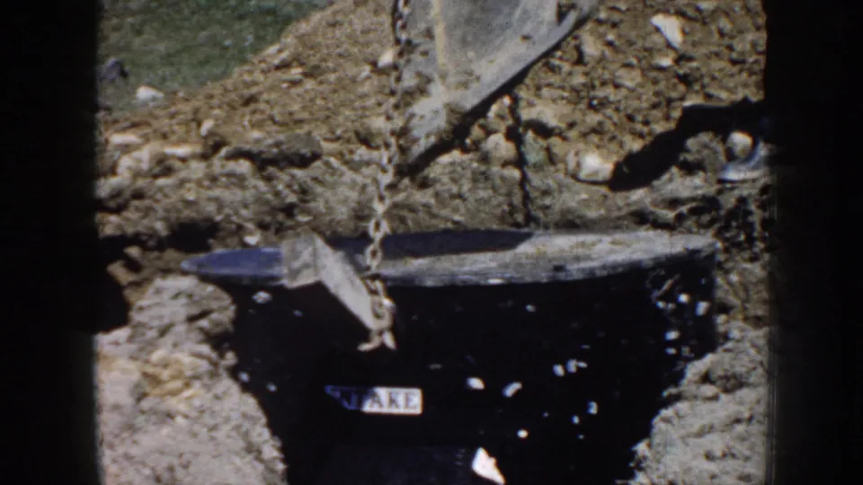 1958 Nebraska 8mm frame showing a black utility-like cylinder in a dug trench, suspended by a chain from an excavator bucket.