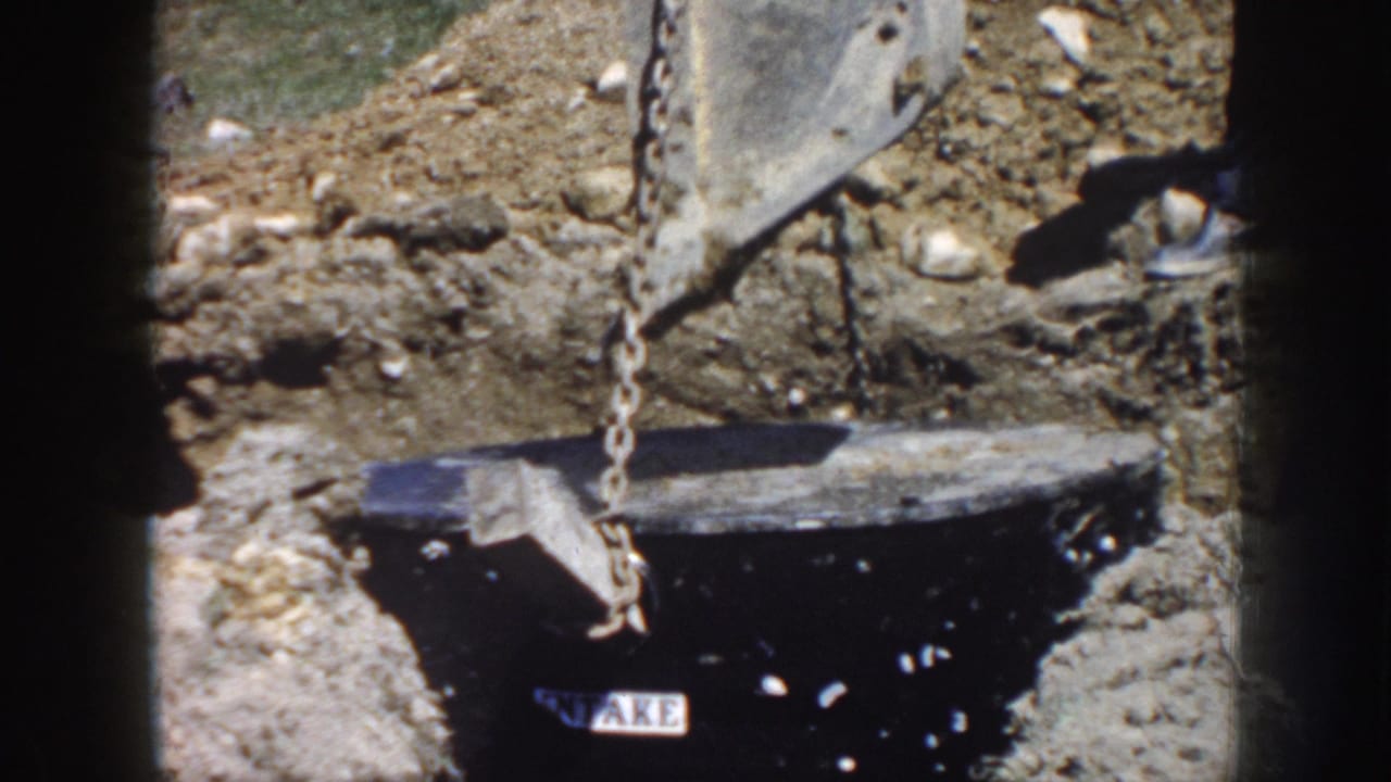 Excavator bucket and chain lowering a black cylinder into disturbed earth.