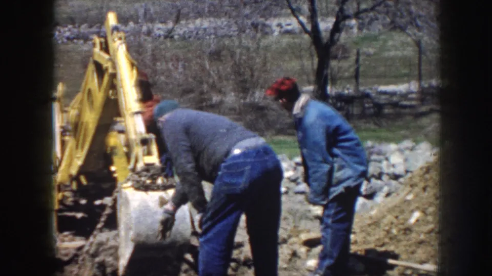 Three men coordinate around a yellow excavator in a rural Nebraska worksite, with bare trees and a stone wall behind them.