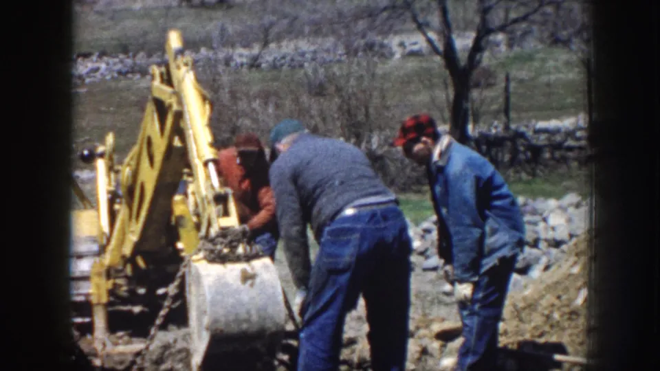 Rural Nebraska 1958 frame with three men working beside a yellow excavator, disturbed soil, bare trees, and a low stone wall.