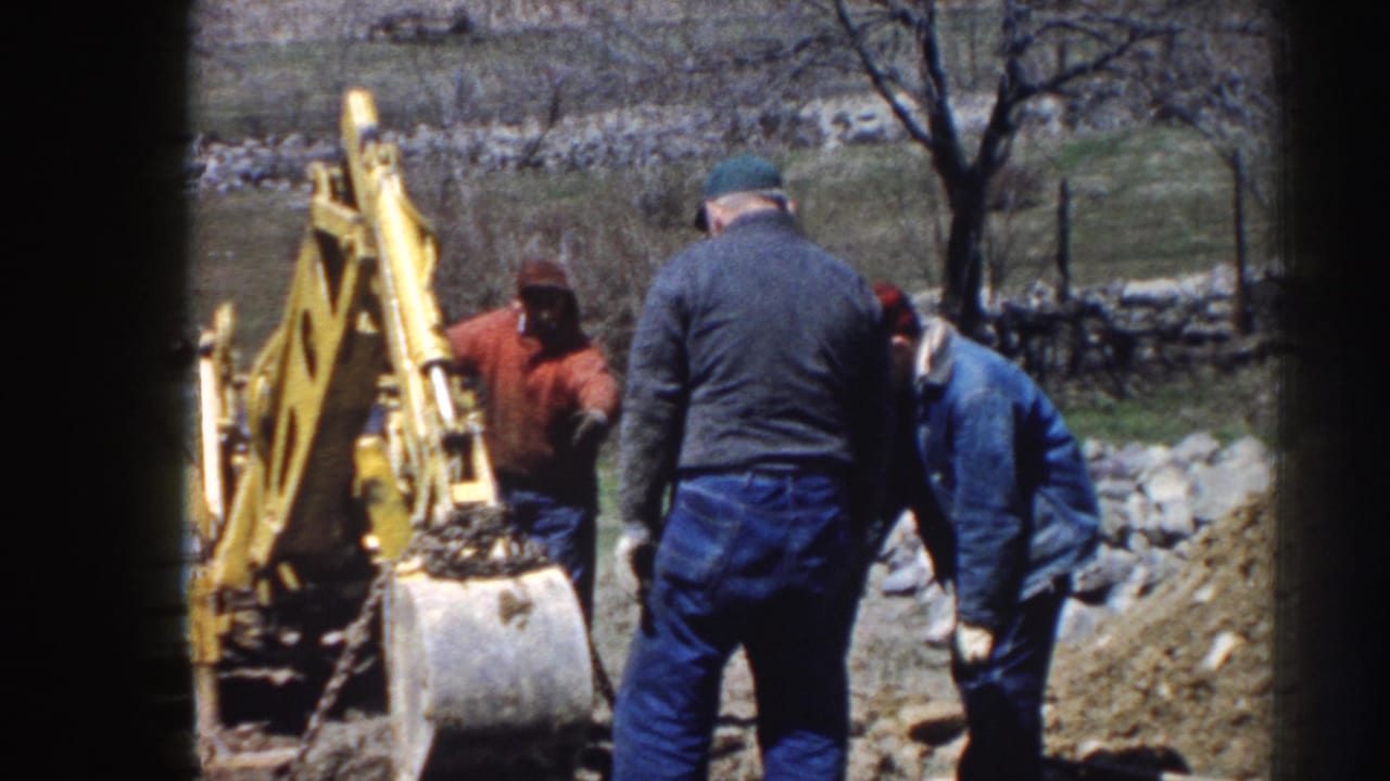 1958 Nebraska home-movie frame showing three adult men working beside a yellow backhoe, with rocks and bare trees behind them.