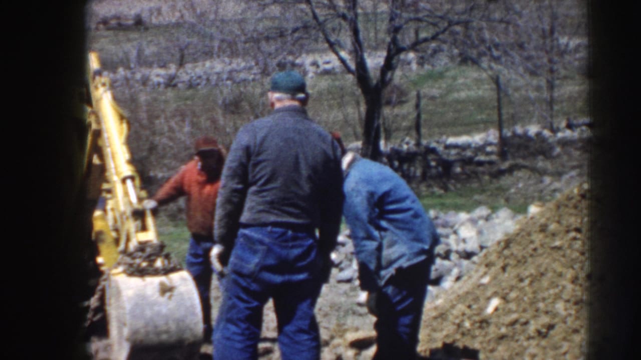 Men in work clothes stand beside a yellow backhoe near a soil pile.