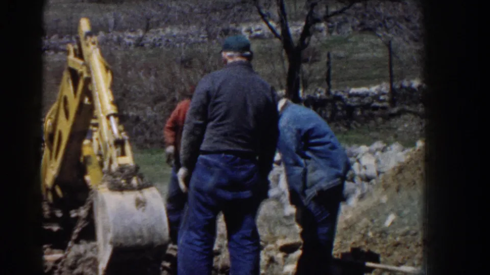 1958 Nebraska home-movie frame showing men beside a yellow backhoe bucket, with rocks, a dirt pile, and bare trees in the background.