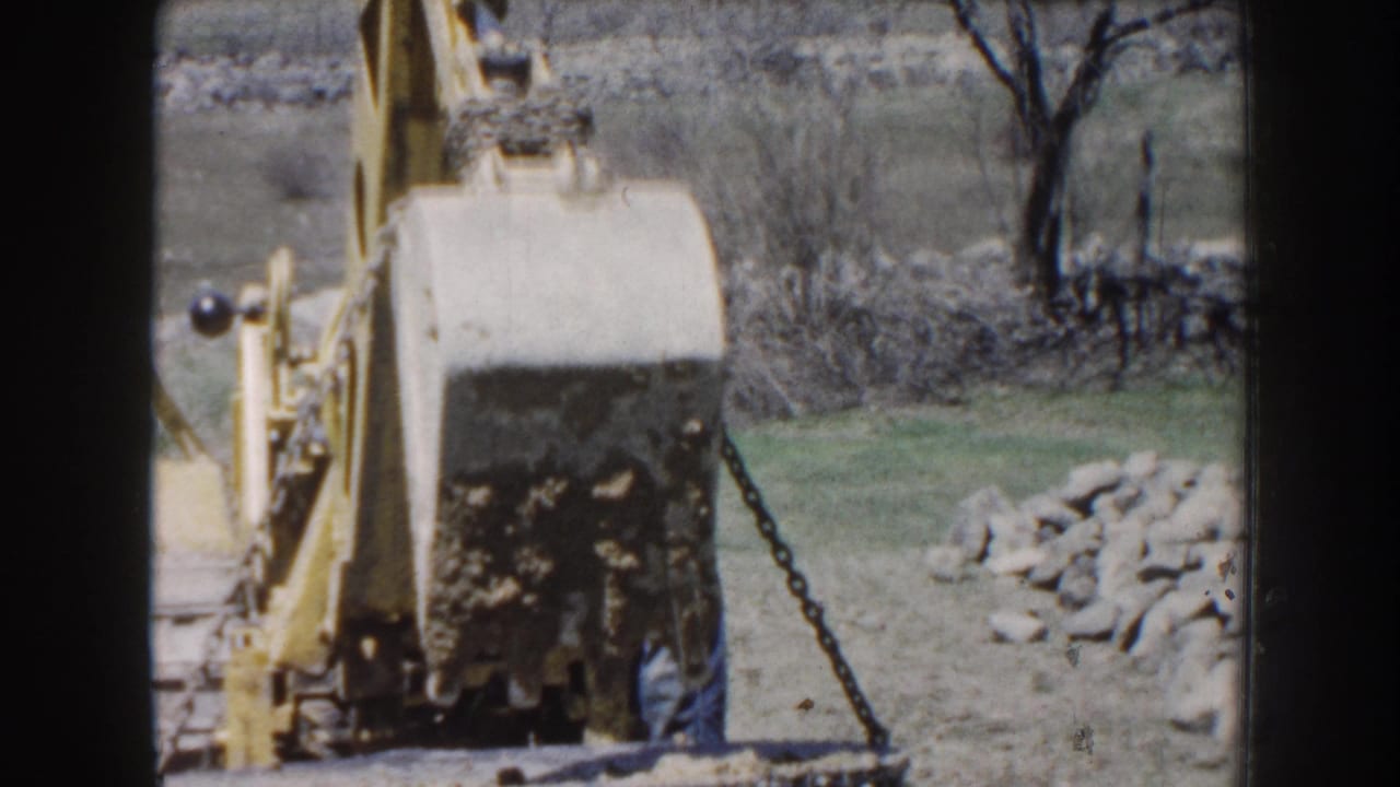 1959 Nebraska 8mm frame showing a yellow excavator beginning to lift a dirt-covered cylindrical concrete object in a rural setting.