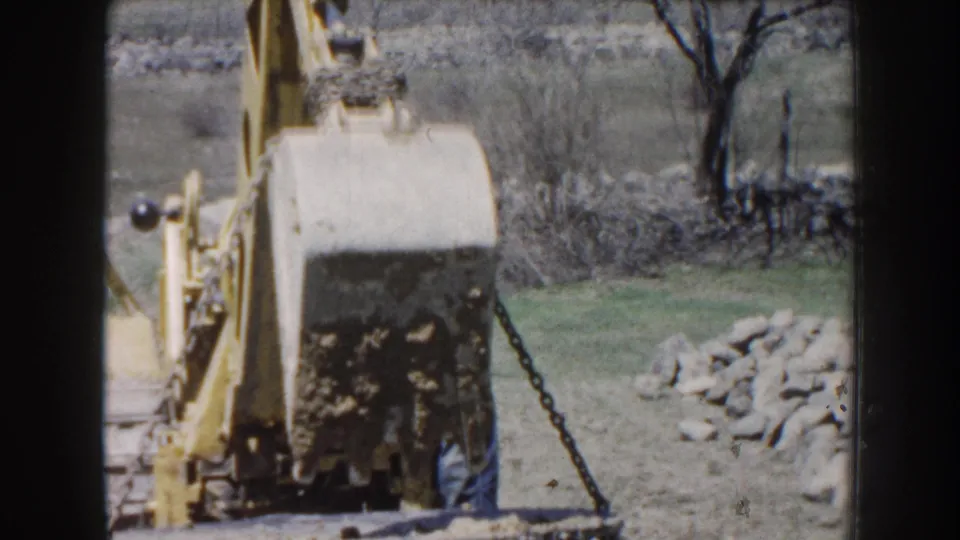 1959 Nebraska 8mm frame showing a yellow excavator beginning to lift a dirt-covered cylindrical concrete object in a rural setting.