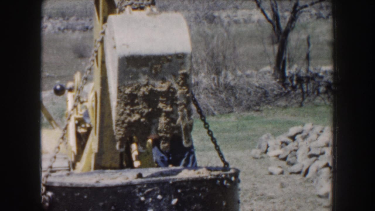Concrete cylinder suspended by chains beside a yellow excavator in a grassy rural area.