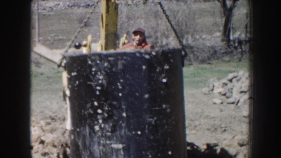 The lifted concrete cylinder fills the foreground while a worker is partly visible behind the excavator equipment in the 1959 Nebraska footage.