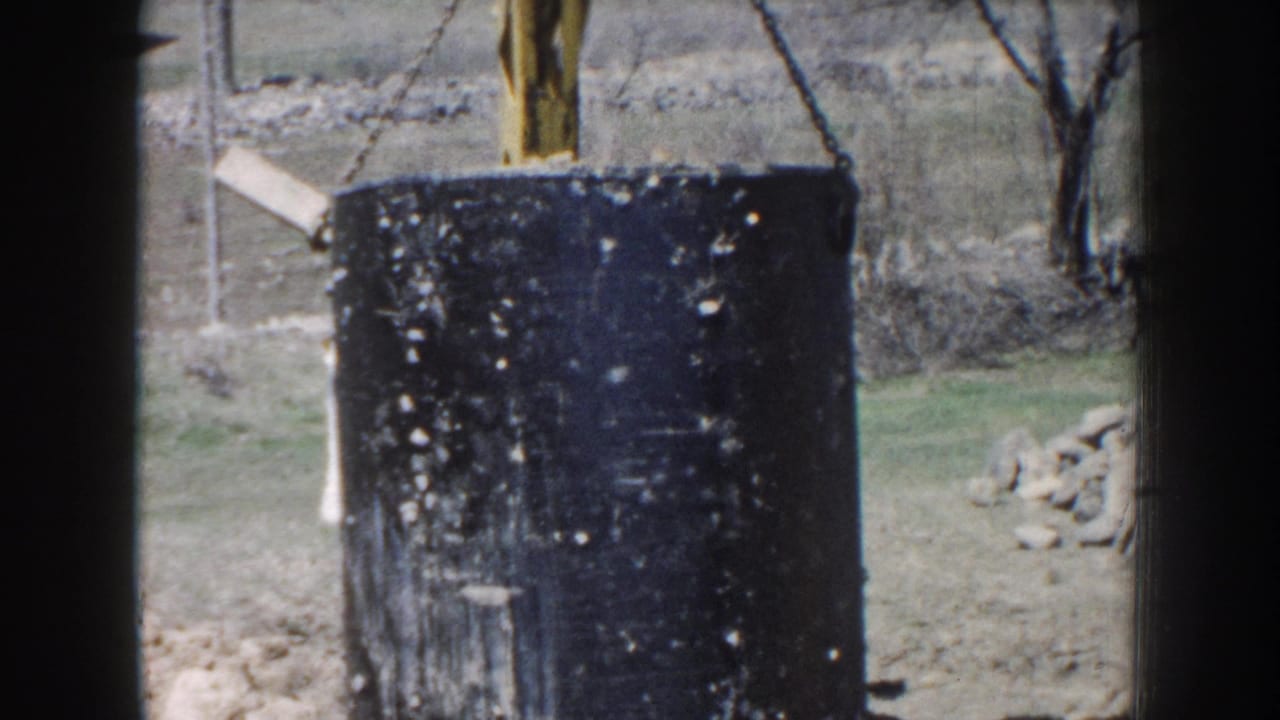 Large black tank suspended near a yellow excavator with rocks and trees behind it.