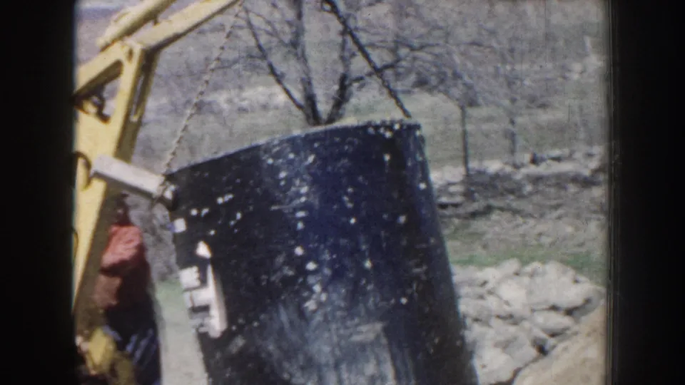 1959 Nebraska home-movie frame showing a worker near a tilted black septic tank as yellow excavation equipment lifts it.