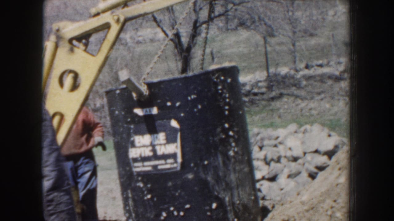 1959 Nebraska home-movie frame showing a crane hoisting a black Empire Septic Tank as a man in a cap observes near a stone wall.