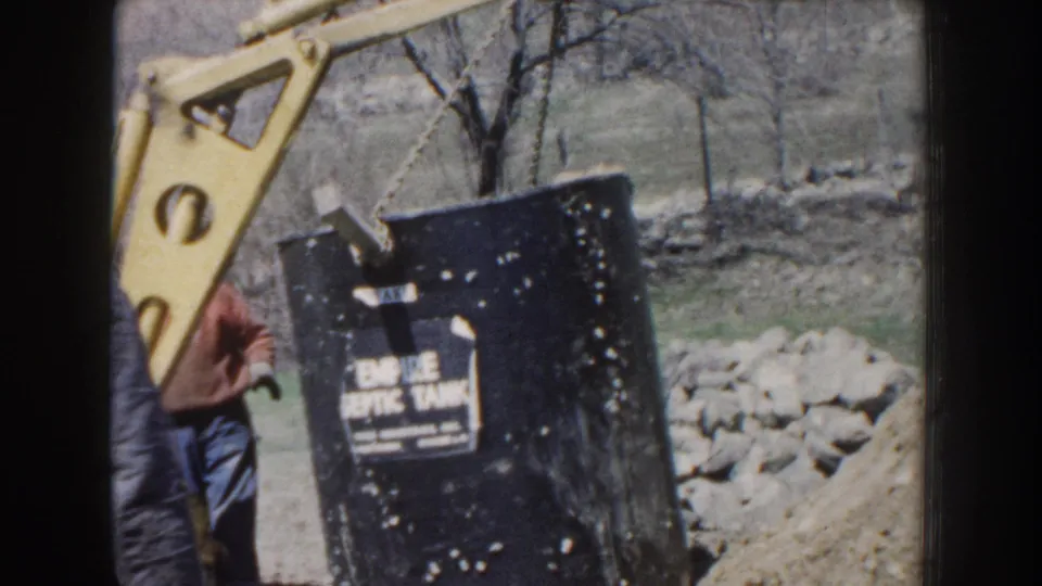 1959 Nebraska home-movie frame showing a crane hoisting a black Empire Septic Tank as a man in a cap observes near a stone wall.