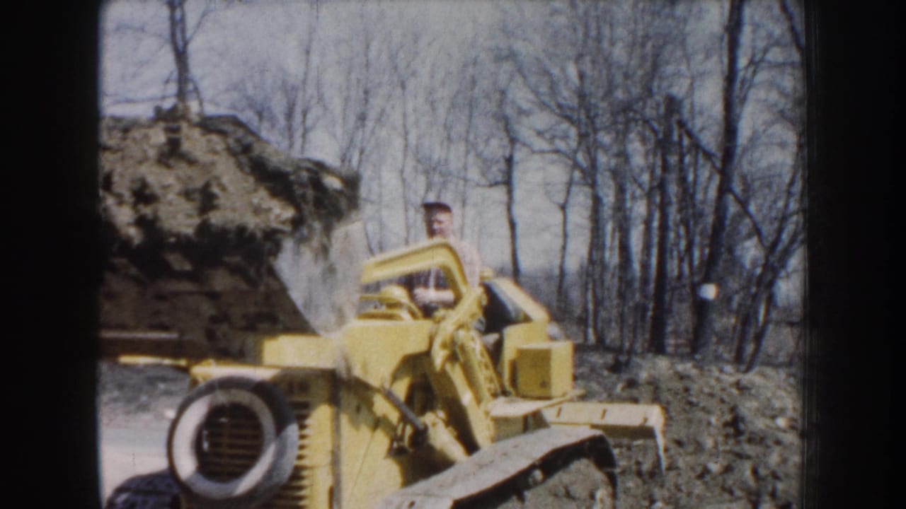 Yellow front-end loader carries a bucket of dirt near bare trees.