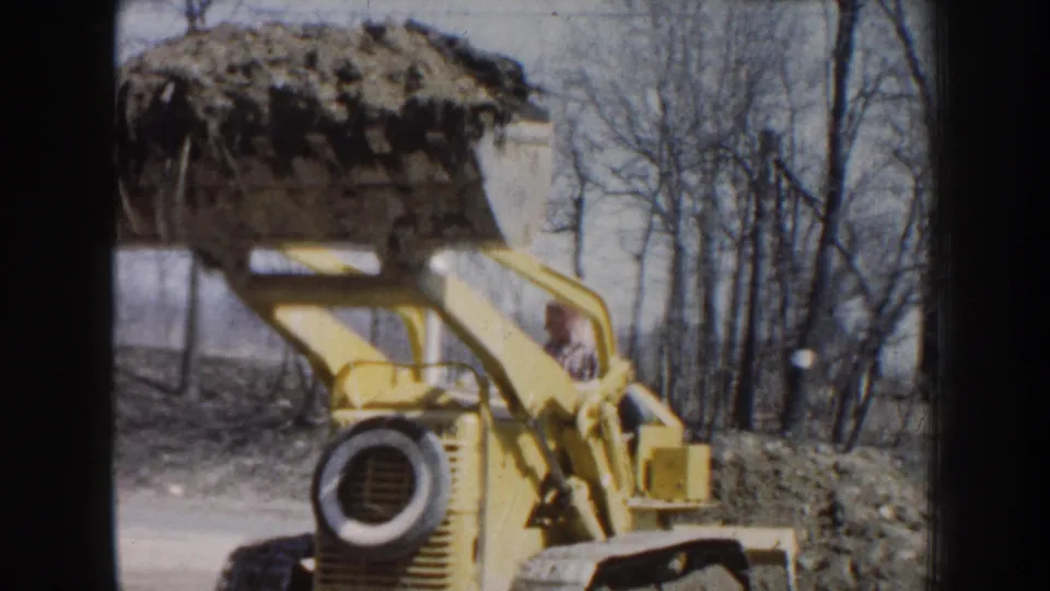 1959 Nebraska home-movie frame showing the front of a yellow loader with a raised bucket carrying soil at a rural outdoor worksite.
