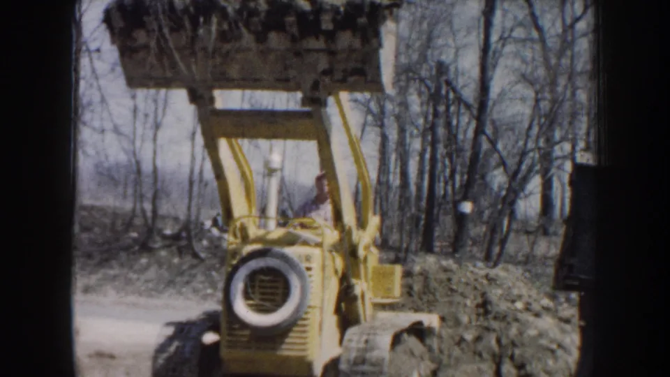 1959 Nebraska home-movie frame showing a yellow loader facing the camera with its bucket raised high against a line of bare trees.