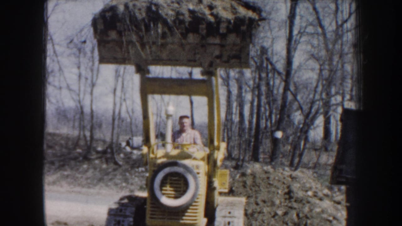 1959 Nebraska home-movie frame showing a man driving a yellow front-end loader with its bucket raised near bare trees.
