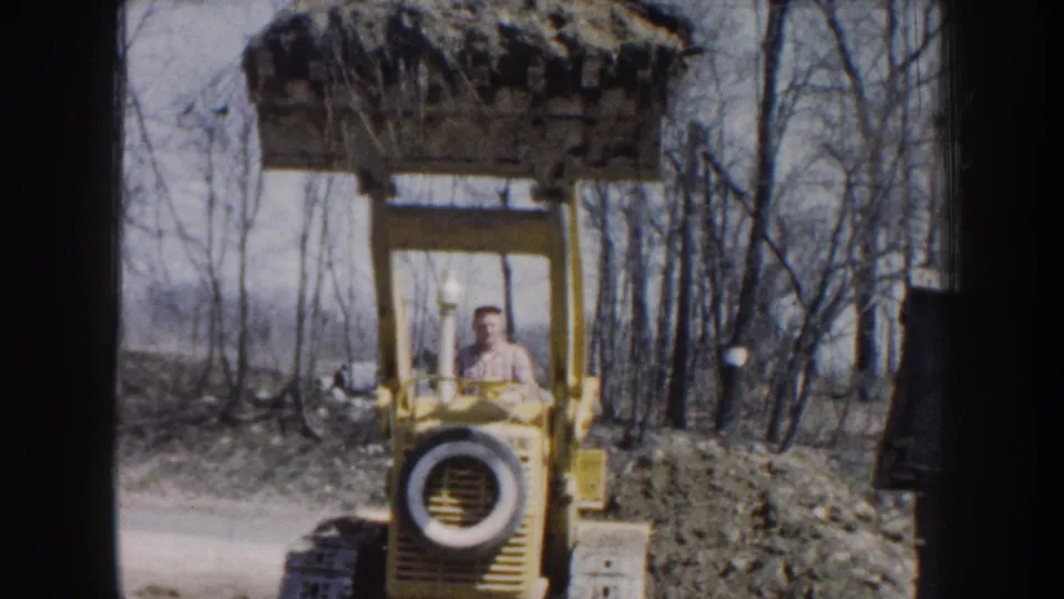1959 Nebraska home-movie frame showing a man driving a yellow front-end loader with its bucket raised near bare trees.