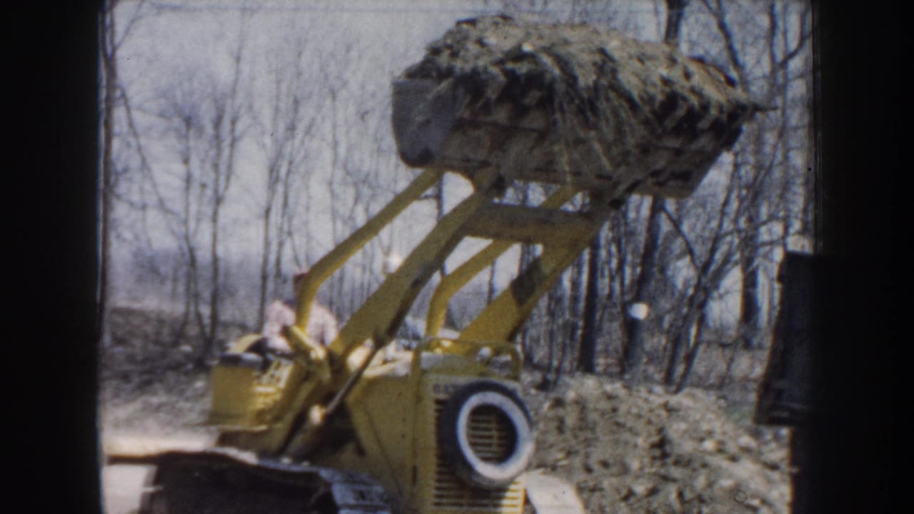 Yellow loader carries a raised bucket of dirt or brush beside bare trees.