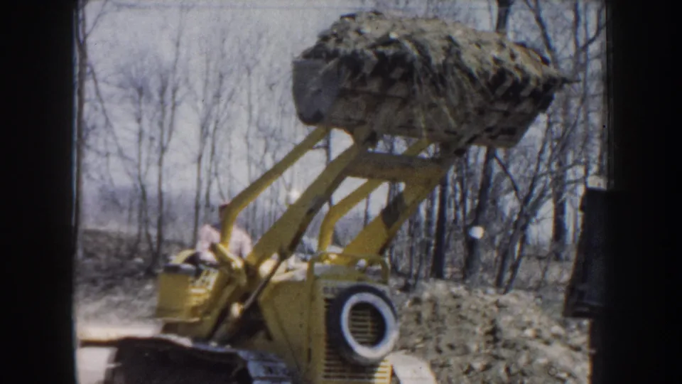 The yellow front-end loader moves through a Nebraska wooded area with a raised bucket holding soil and debris.