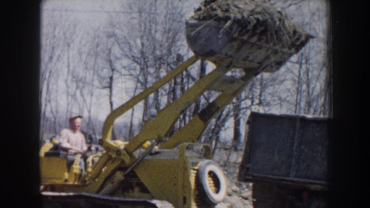 In Nebraska 1959 home-movie footage, an adult operator drives a yellow loader with a raised dirt-filled bucket beside a truck bed.