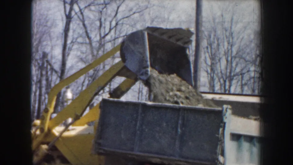The front-end loader’s bucket remains tipped over the truck bed as dirt falls during the 1959 Nebraska earthmoving sequence.
