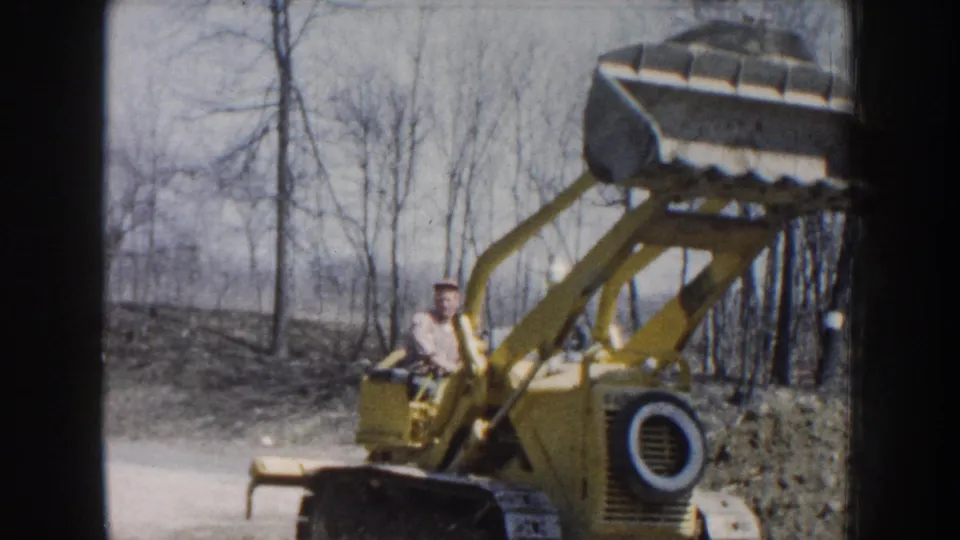 An adult operator sits on the yellow loader as the bucket is raised or clearing the truck after dumping dirt in a wooded Nebraska work area.