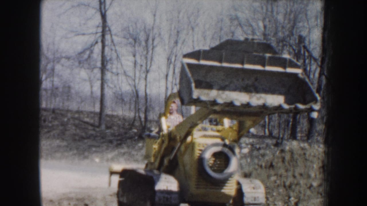 1959 Nebraska 8mm frame showing a yellow tracked loader approaching with its front bucket raised near bare trees.