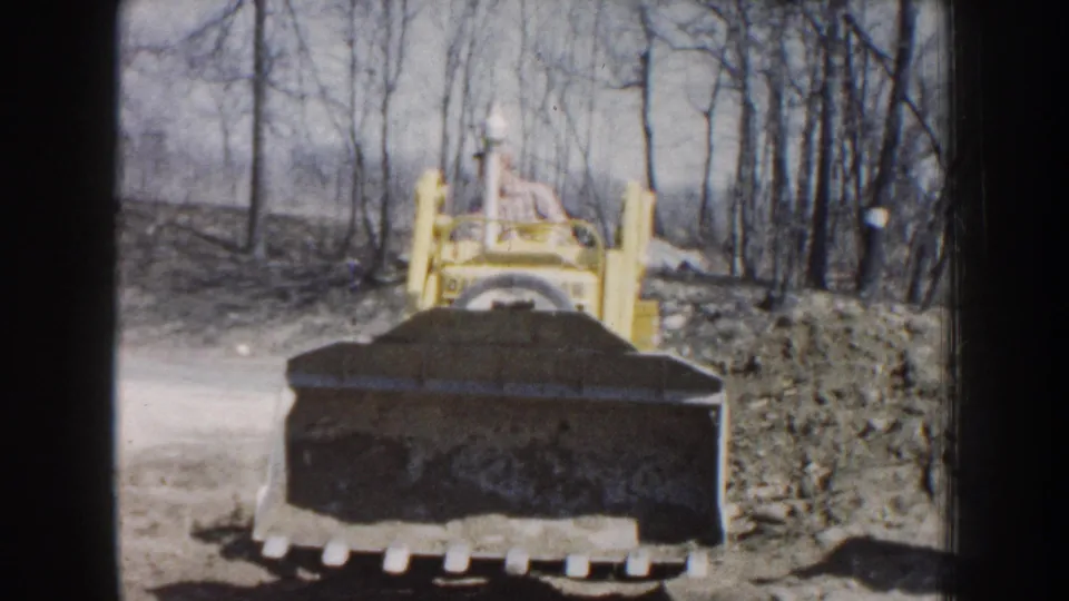 Yellow tracked loader with a dirt-filled bucket in a wooded Nebraska work area, captured on 1959 8mm film.