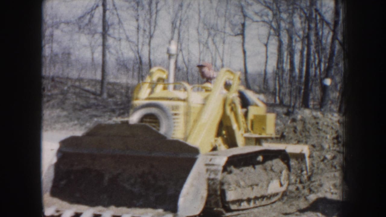 1959 Nebraska 8mm frame showing a man operating a yellow tracked bulldozer in a wooded dirt area.