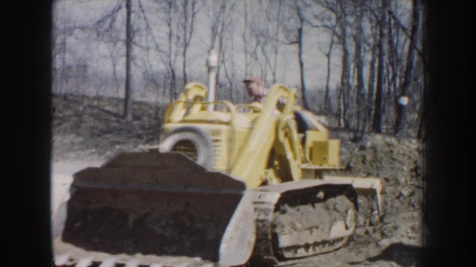 1959 Nebraska 8mm frame showing a man operating a yellow tracked bulldozer in a wooded dirt area.