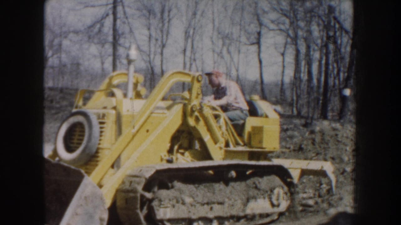1959 Nebraska home-movie frame of a capped man operating a yellow tracked bulldozer near bare trees.