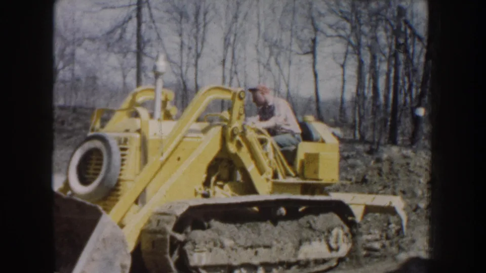 1959 Nebraska home-movie frame of a capped man operating a yellow tracked bulldozer near bare trees.