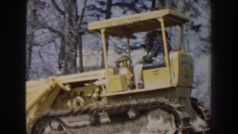 1959 Nebraska frame showing a yellow bulldozer with a canopy and seated operator in a wooded outdoor setting.