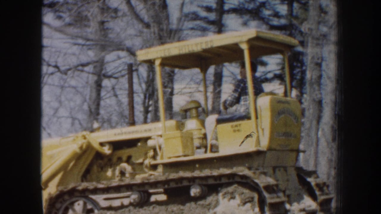 1959 Nebraska 8mm frame showing a yellow Caterpillar bulldozer with a seated operator moving through a wooded dirt area.
