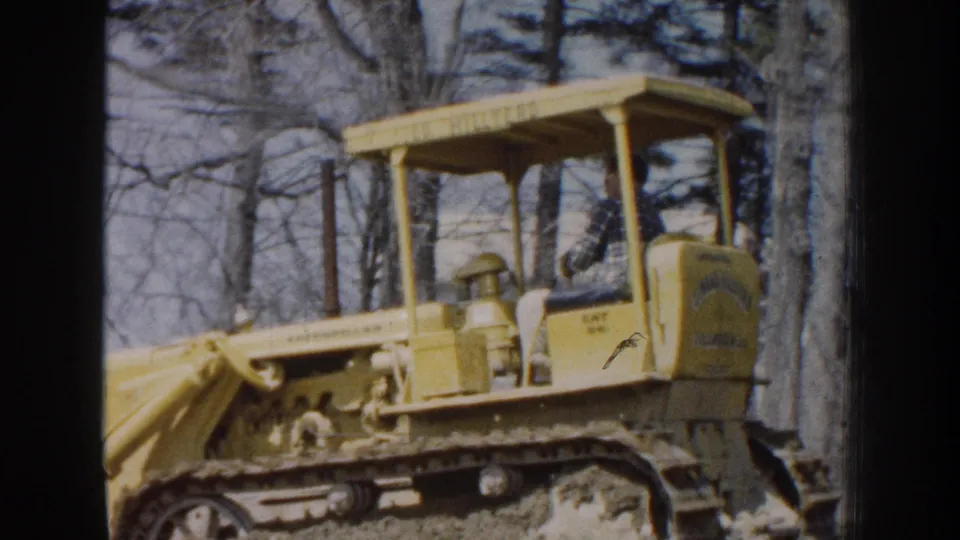 1959 Nebraska 8mm frame showing a yellow Caterpillar bulldozer with a seated operator moving through a wooded dirt area.