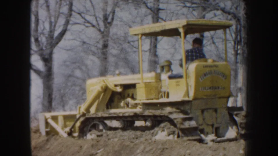 1959 Nebraska 8mm frame showing the side of a yellow bulldozer and operator, with the blade set low near bare trees.