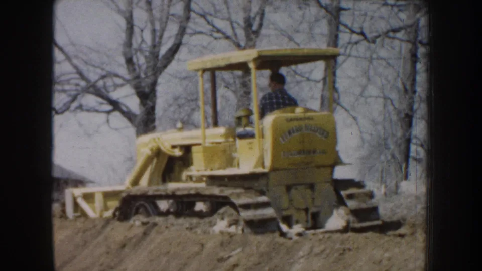 1959 Nebraska 8mm frame showing a yellow tracked bulldozer with a seated male operator near bare trees and a dirt embankment.
