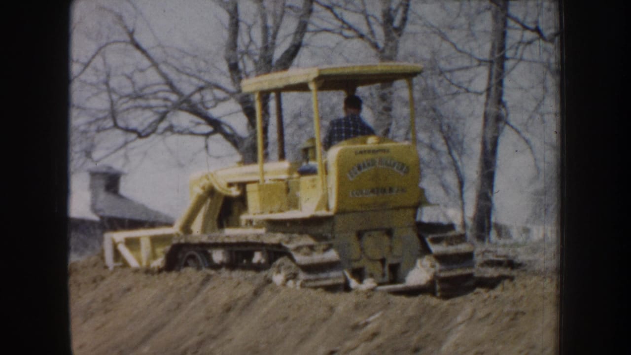 Yellow bulldozer seen from the rear with operator, tracks, blade, and bare trees.