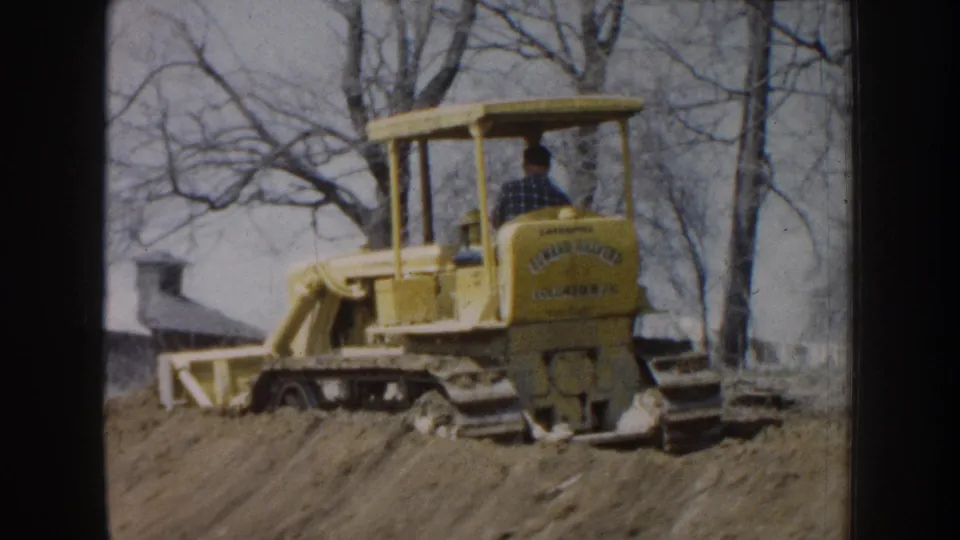 Rear view of a yellow bulldozer in Nebraska, 1959, with a male operator in an open cab and leafless trees behind it.