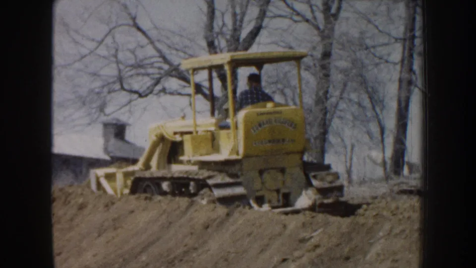 1959 Nebraska home-movie frame of a yellow tracked bulldozer and operator working along a dirt bank in a rural setting.