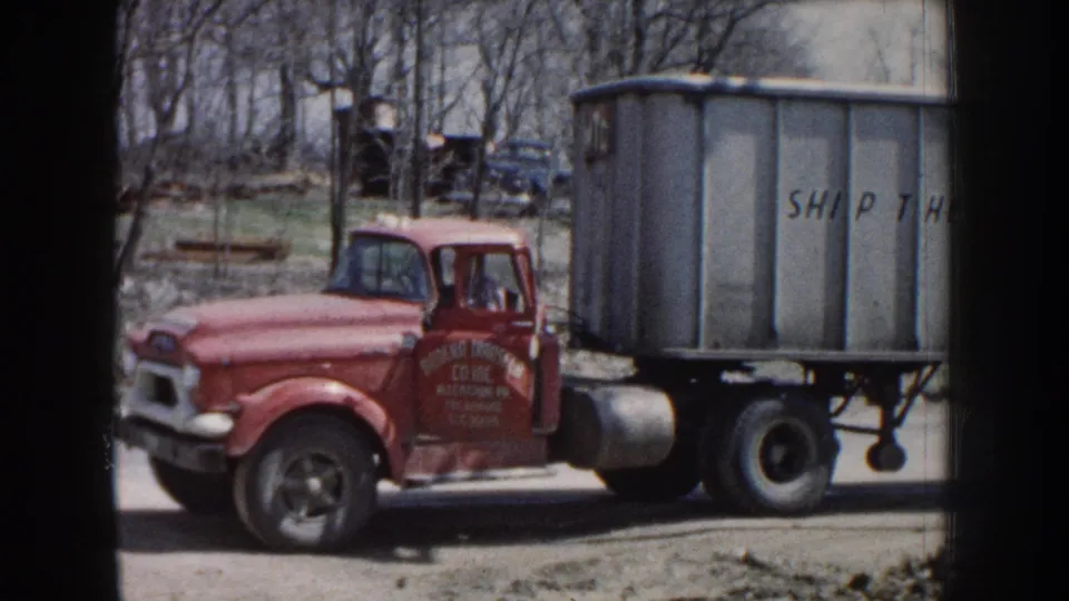 1959 Nebraska 8mm frame showing a red GMC truck beside a gray trailer marked with transport company lettering.