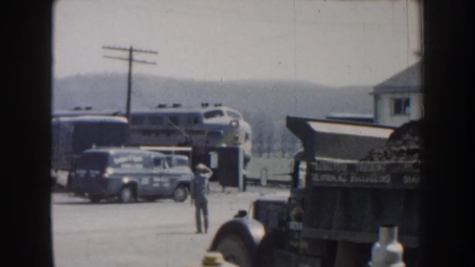 1959 Nebraska station scene with a passenger train arriving behind a van, a standing man, and foreground truck bed.