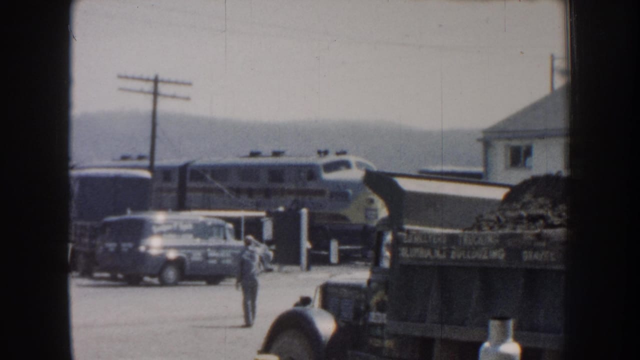 Train passing behind a parked sedan, dump truck, and a man walking across a roadside yard.