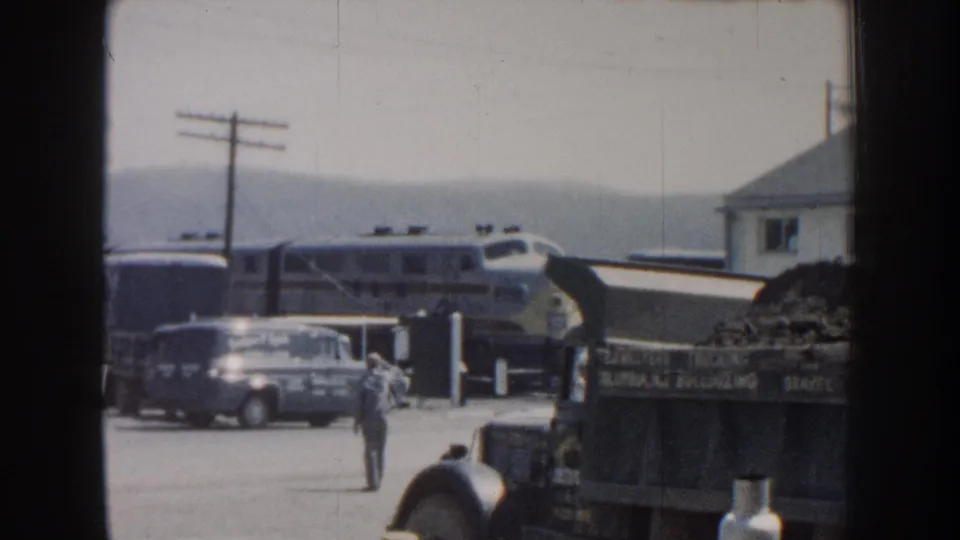 1959 Nebraska home-movie frame showing a passenger train behind parked vehicles as a man walks through a gravel roadside area.
