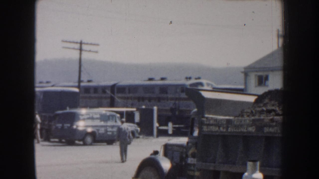 Passenger train, parked car, dump truck, and a pedestrian in a bright roadside yard.