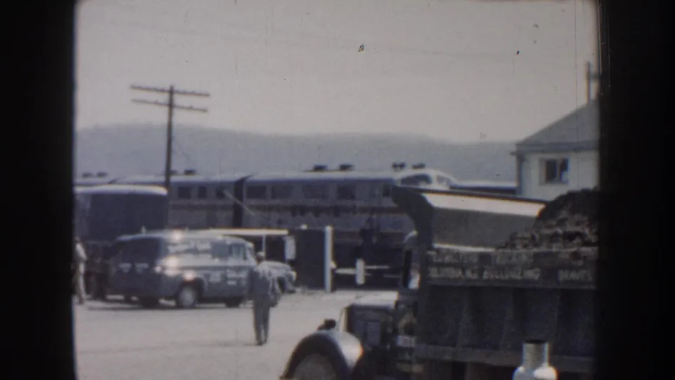A Nebraska rail crossing scene with a streamlined passenger train, mid-century vehicles, and a pedestrian in the foreground.