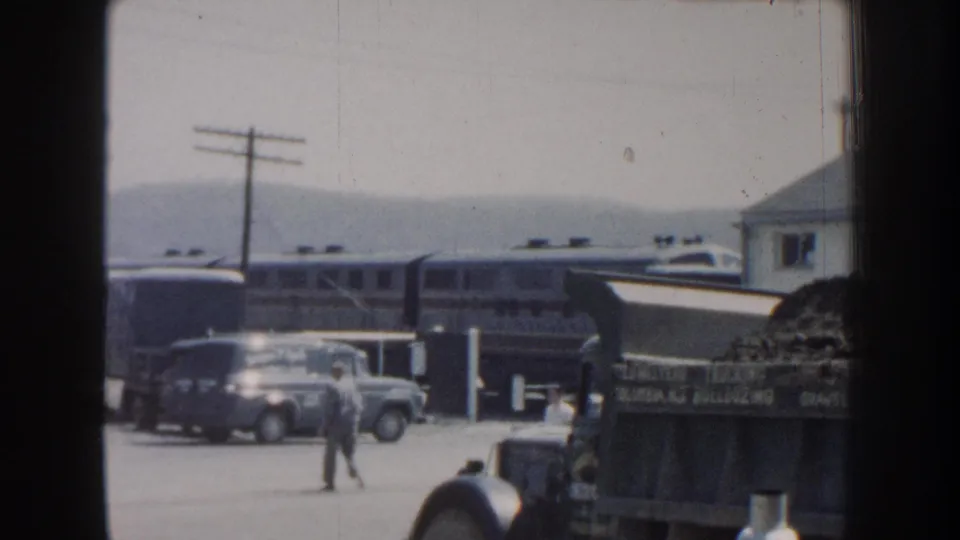 Home-movie view of a man crossing a Nebraska roadside yard while a passenger train and period vehicles fill the background.