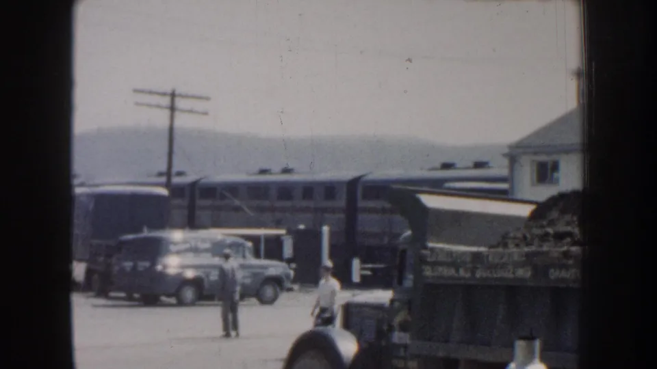 Two visible adults stand or walk near parked vehicles as a passenger train passes through the Nebraska roadside scene.