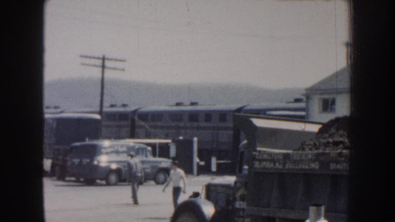1959 Nebraska 8mm frame showing a passenger train, rounded mid-century cars, utility poles, and a foreground truck bed.