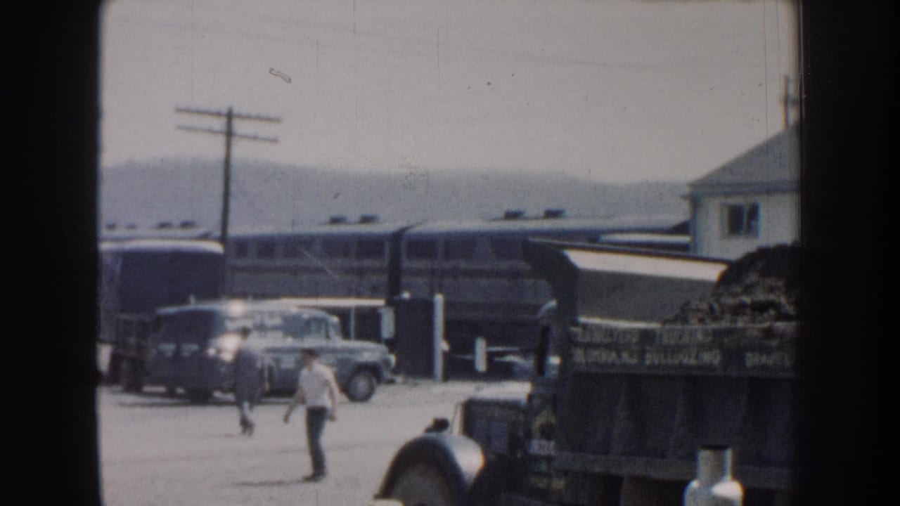1959 Nebraska home-movie frame with a man crossing a rail-side yard near vintage cars, a train, and a loaded truck.